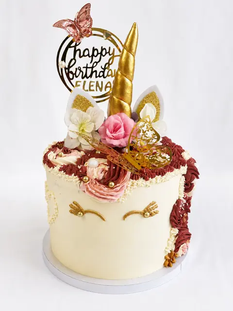 Gâteau d’anniversaire ivoire pour Elena sur le thème de la licorne, décoré d’une corne et d’oreilles dorées, de cils pailletés, de fleurs roses et blanches et d’un papillon